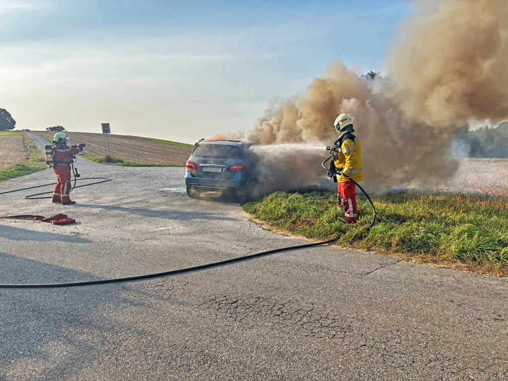 Auch ein Übergreifen der Flammen auf ein angrenzendes Stoppelfeld konnte verhindert werden. Zwei Feuerwehrleute löschen einen silberfarbenen Mercedes, der in Brand steht.